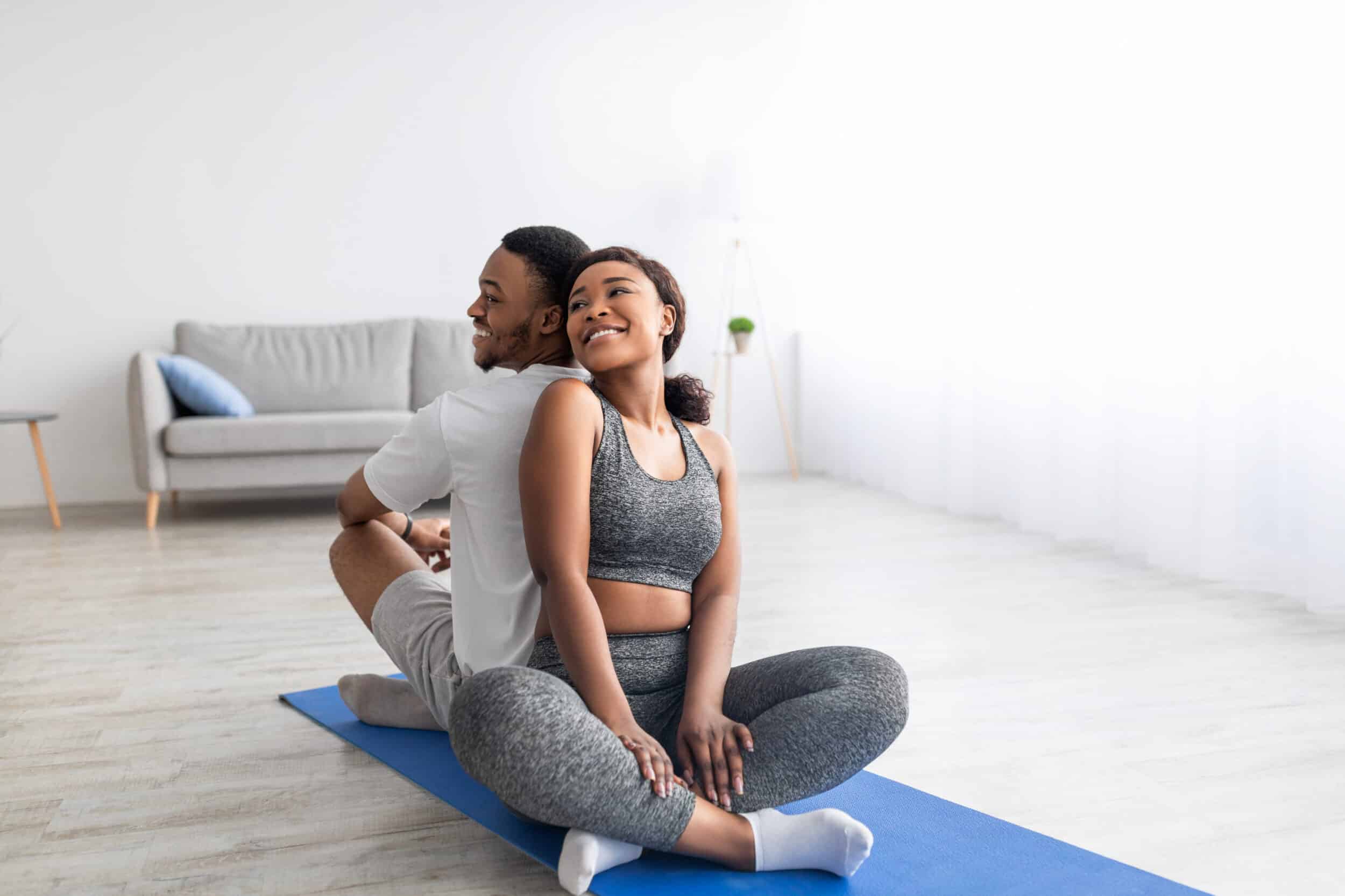 Black lady sitting back to back with her sporty boyfriend in lotus position, having domestic partner yoga practice